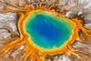 An aerial photo showing a large pool of water that's bright blue in the middle, surrounded by a yellow area and rusty orange zone on the outside. A tiny footpath and people near this spring show it must be at least 100m across.