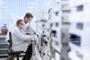 A photo of two white male scientists in labcoats sitting. One of them is sitting on a chair, the other standing next to him. Both are intently staring at a screen sitting on a lab bench.