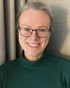 Portrait photo of Helen Sneddon, smiling into the camera, standing in front of some beige curtains