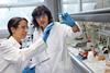 Two women wearing white lab coats, goggles and blue nitrile gloves look at a vial being held up by the person on the right. They are stood in a lab next to a fume hood